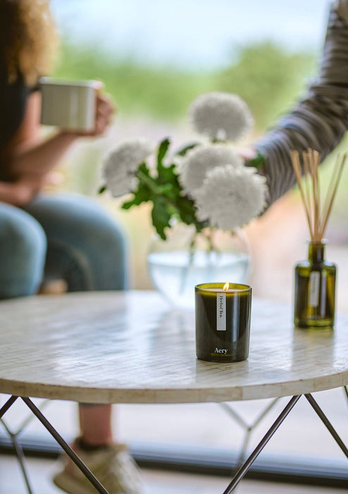 Green Herbal Tea candle by Aery displayed next to diffuser and vase of white flowers sat on cream white table  