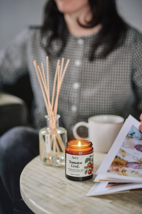 Tomato Leaf jar candle by Aery sat on circle table with diffuser and person reading book