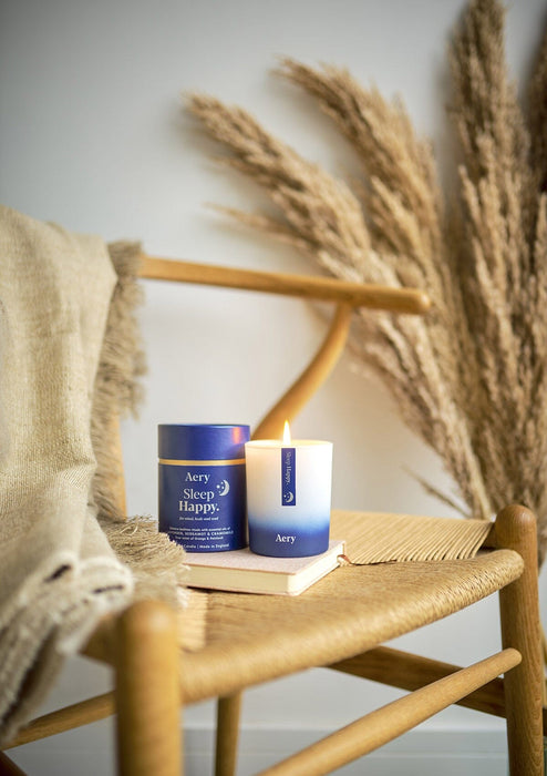 Blue Sleep Happy candle by aery displayed next to product packaging sat on wicker chair 