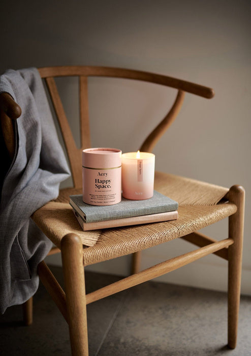 Pink happy space candle by Aery displayed next to product packaging sat on wicker chair 